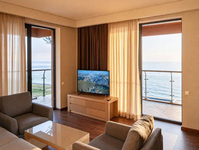 One Bedroom Sea Front & Batumi Side — Living room with mounted TV and twin balcony doors framing the sea
